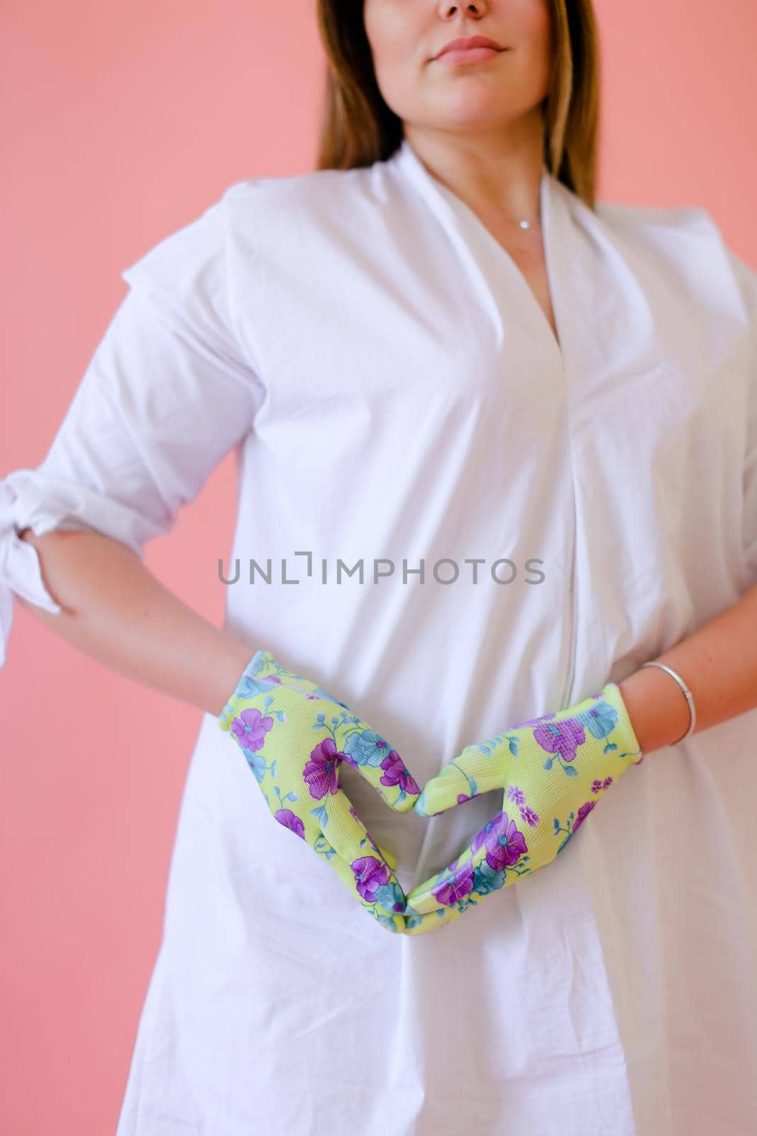 Female person wearing white dress showing heart gesture on belly in pink monophonic background. Concept of pregnant photo session.