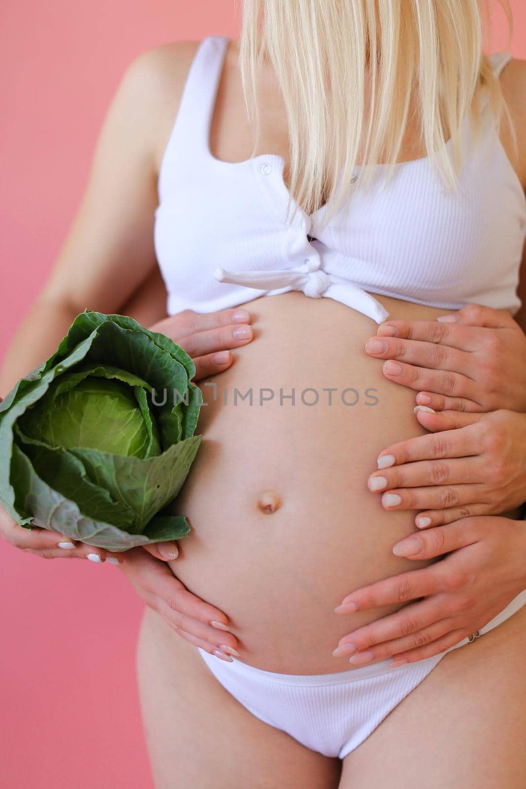 Closeup pregnant woman in underwear keeping cabbage in pink monophonic background. Concept of expactant photo session.