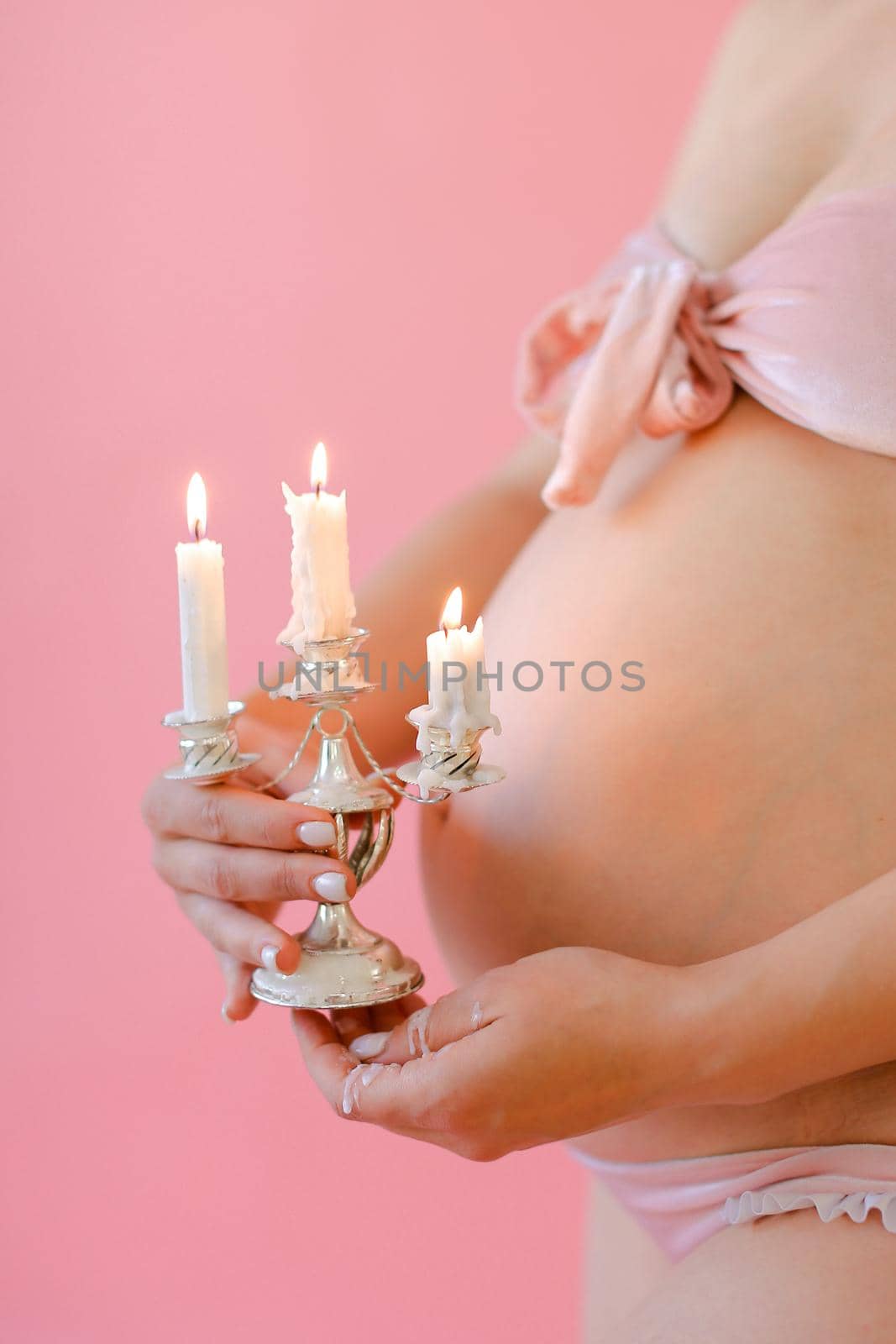 Closeup happy pregnant woman in underwear holding belly and keeping candles in pink monophonic background. Concept of expactant photo session.