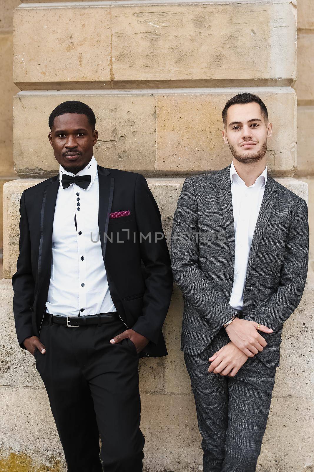 Afro american and caucasian smiling gays standing near building and wearing suits. Concept of lgbt and walking in city.