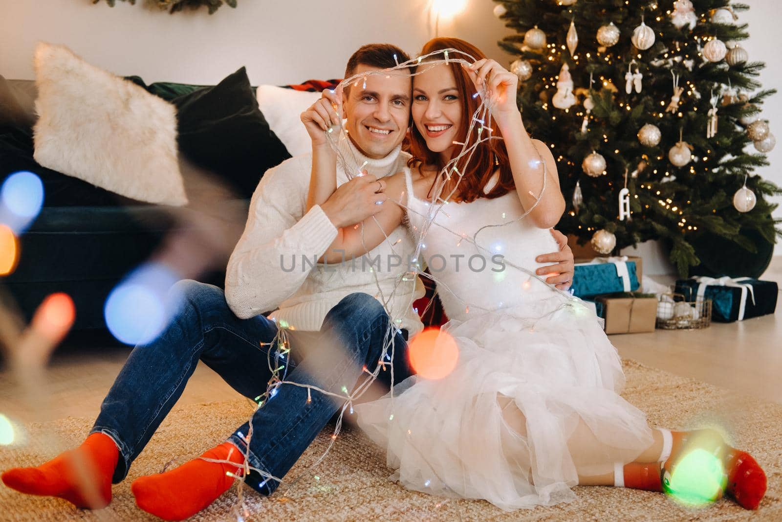 a happy married couple is sitting at home on the floor near the Christmas tree.