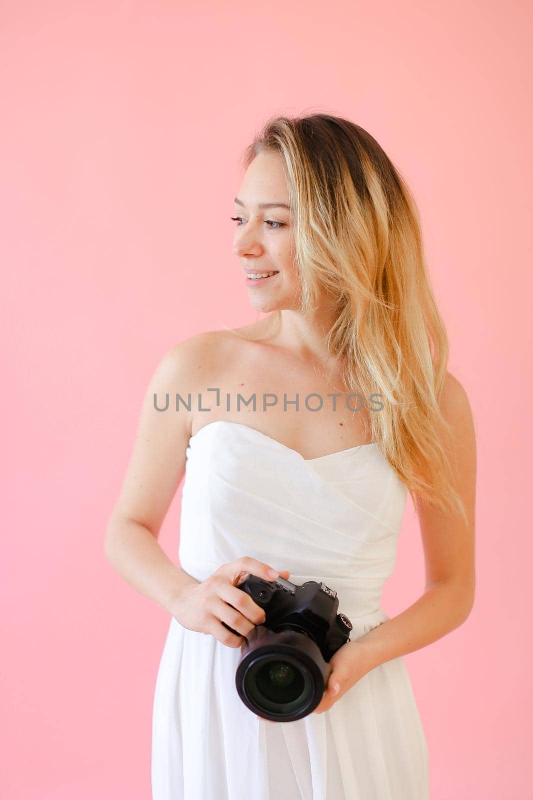 Young gladden female photographer with black camera in pink monophonic background. Concept of modern profession and photo session.