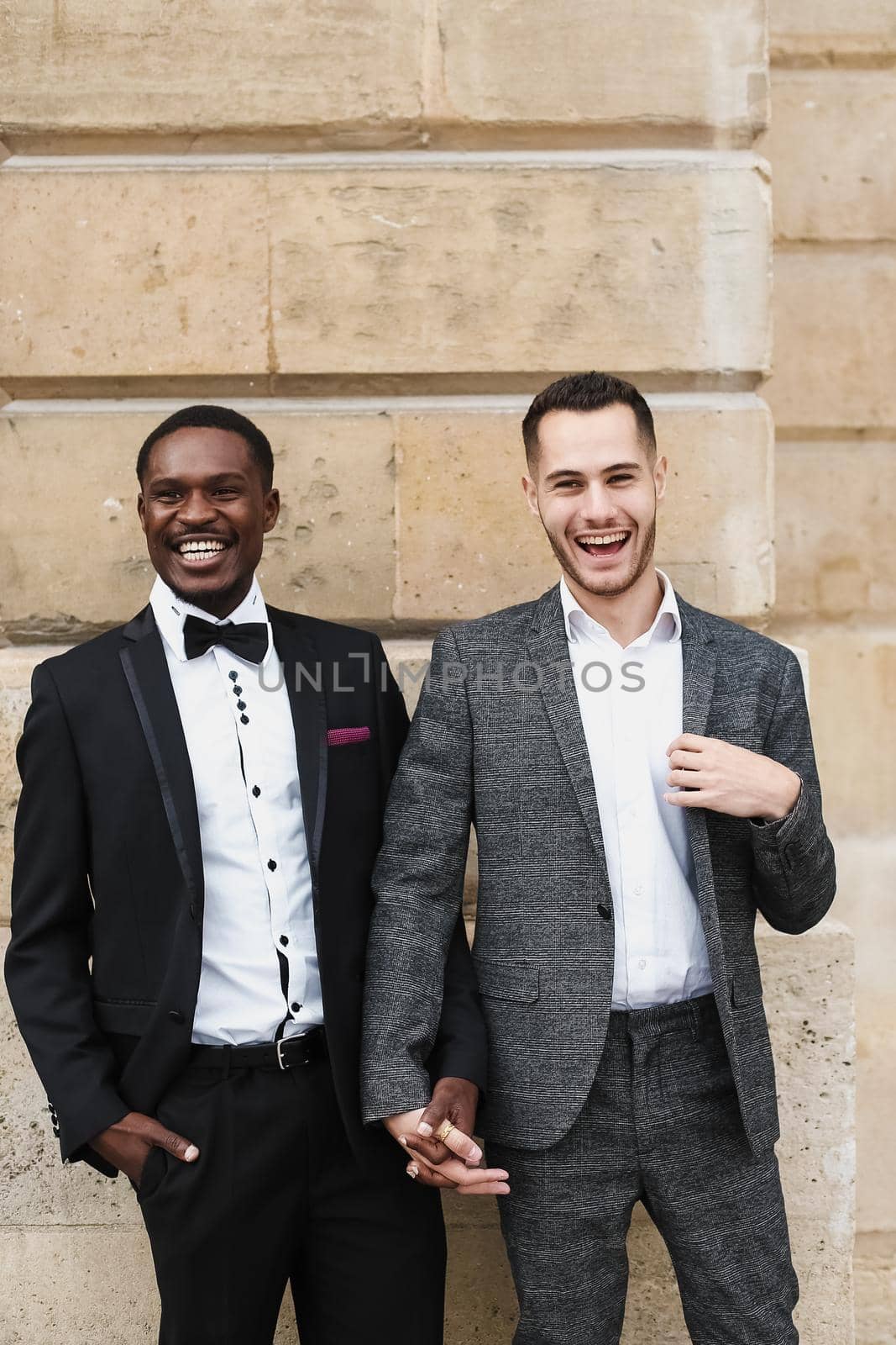 Afro american and european gays standing near building and wearing suits. Concept of lgbt and walking in city.
