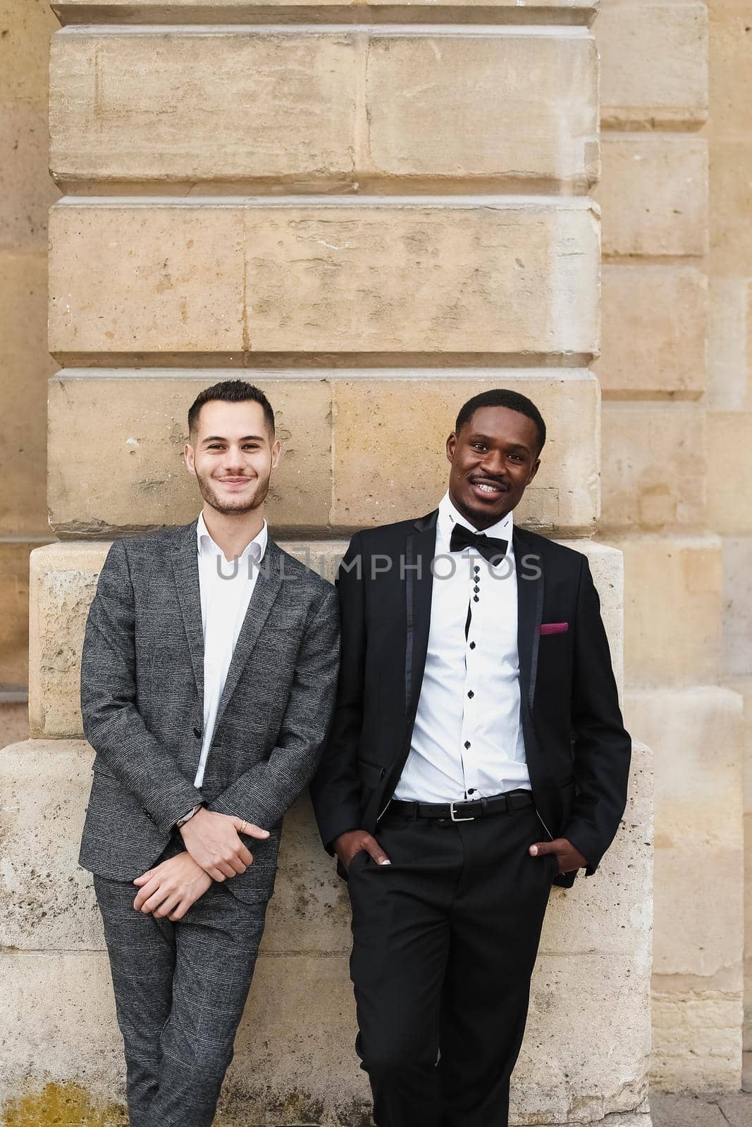 Afro american and european gays standing near building and wearing suits. Concept of lgbt and walking in city.