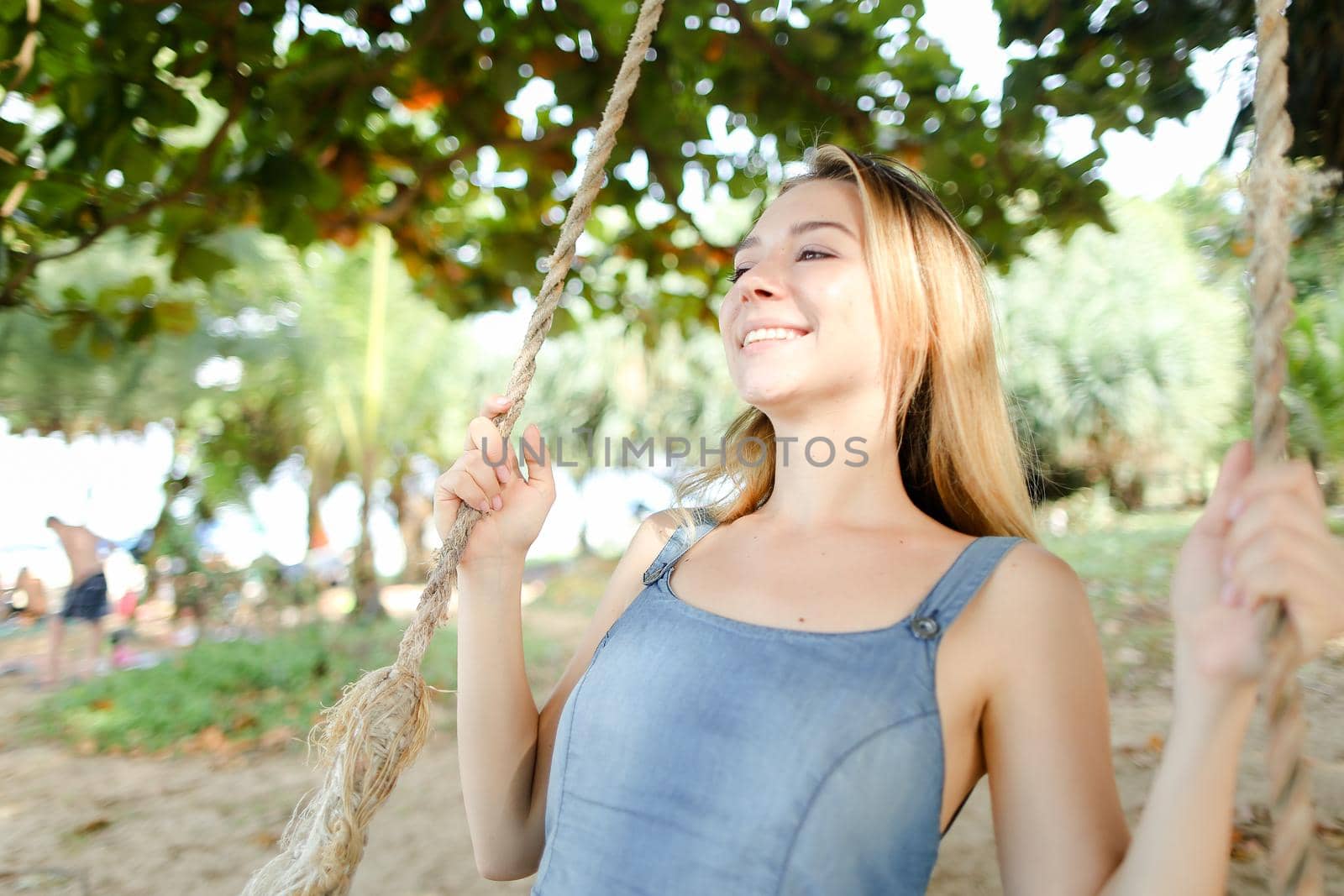 Young smiling girl riding on swing and wearing jeans sundress, sand in background. Concept of summer vacations and leisure time.