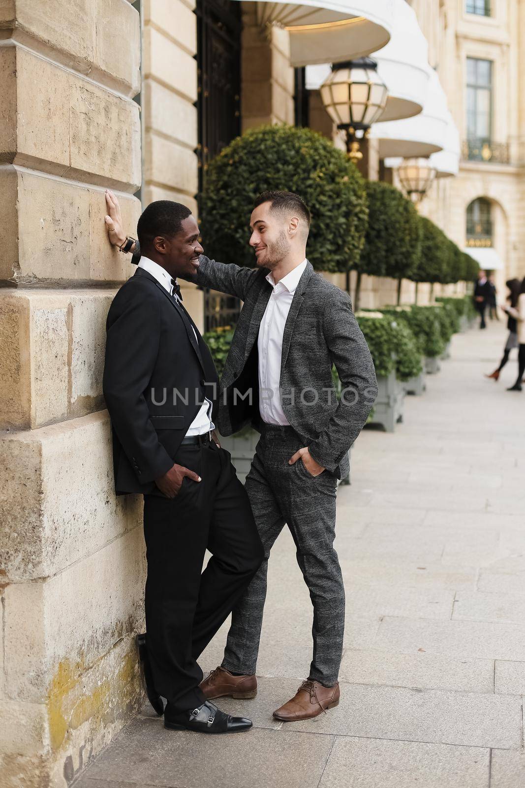 Afro american and european gays standing near building and wearing suits and talking outside. Concept of lgbt and walking in city.