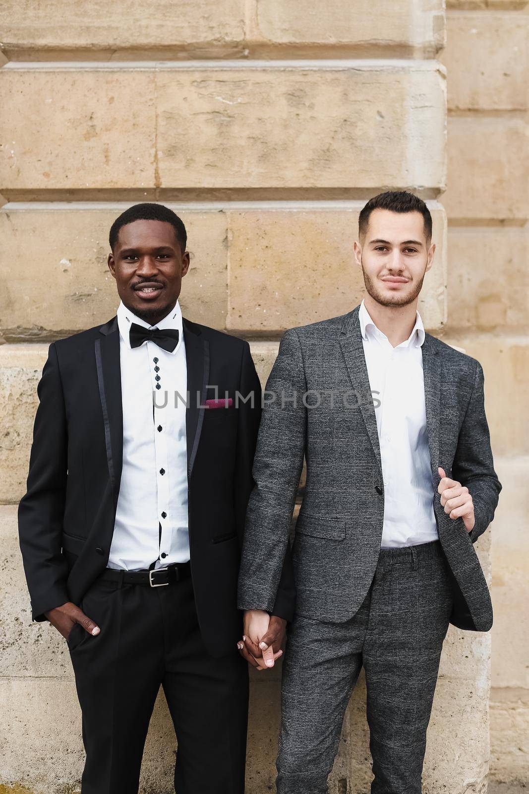 Afro american and caucasian smiling gays standing near building and wearing suits. Concept of lgbt and walking in city.