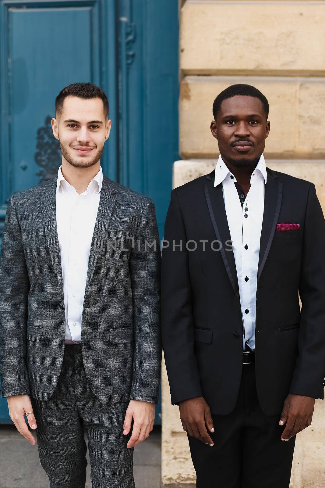 Two men, caucasian and afro american, wearing suits standing near building. by sisterspro