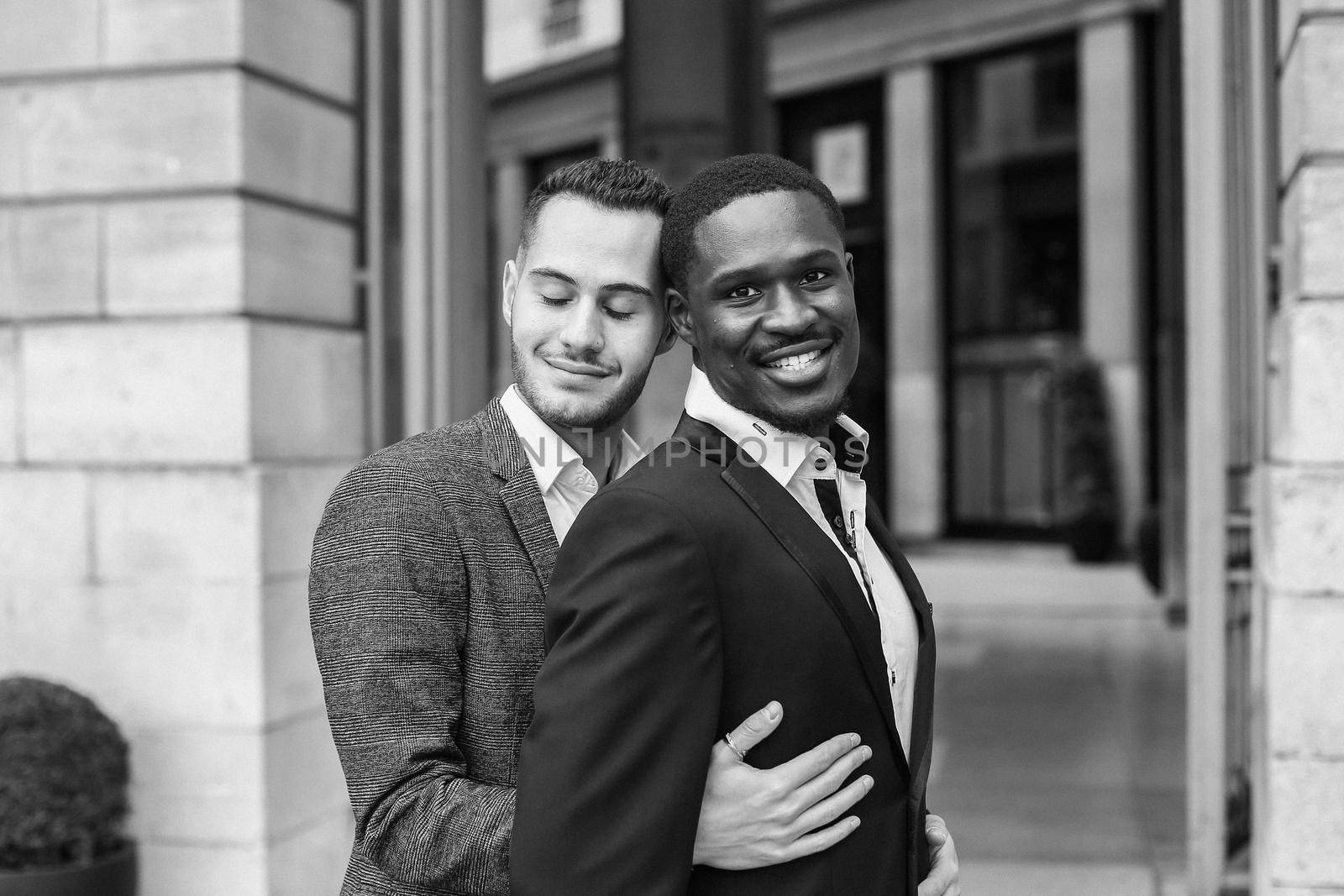 Black and white bw portrait Caucasian man hugging afro american guy outside and wearing suit. Concept of happy same sex couple and gays in Paris
