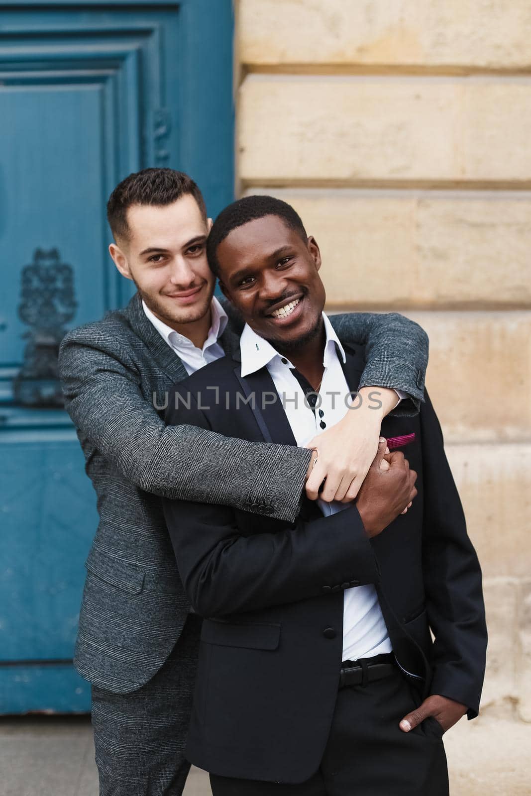 Two boys, caucasian and afro american, wearing suits standing near building and hugging. Concept of gays and lgbt.