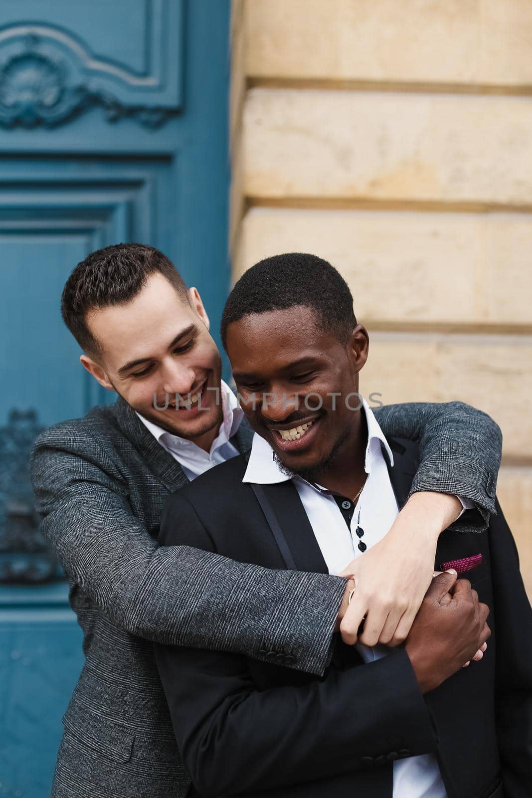 Two men, caucasian and afro american, wearing suits standing near building and hugging. same sex couple by sisterspro