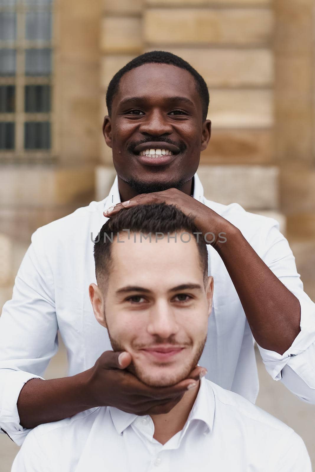 Afro american boy holding caucasian guy head by hands, wearing white shirt. Concept of stylish boy and haircut. Same sex couple, lgbt gays