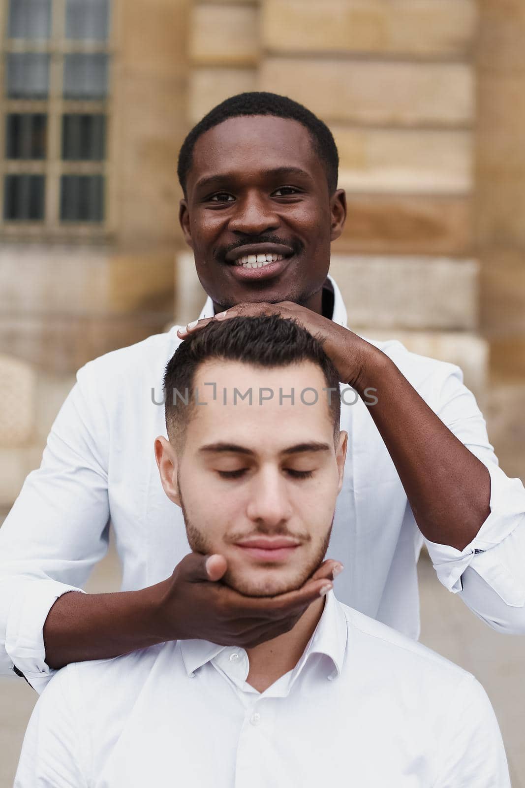 Afro american boy holding caucasian guy head by hands, wearing white shirt. Concept of stylish boy and haircut. Same sex couple, lgbt gays