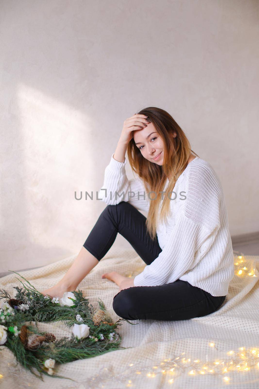Young female person sitting on bed near wreath and yellow garlands. Concept of handmade decorations for Christmas and winter holidays.