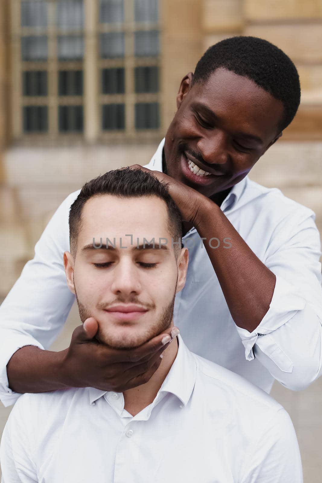 Afro american boy holding caucasian guy head by hands, wearing white shirt. Concept of stylish boy and haircut. Same sex couple, lgbt gays