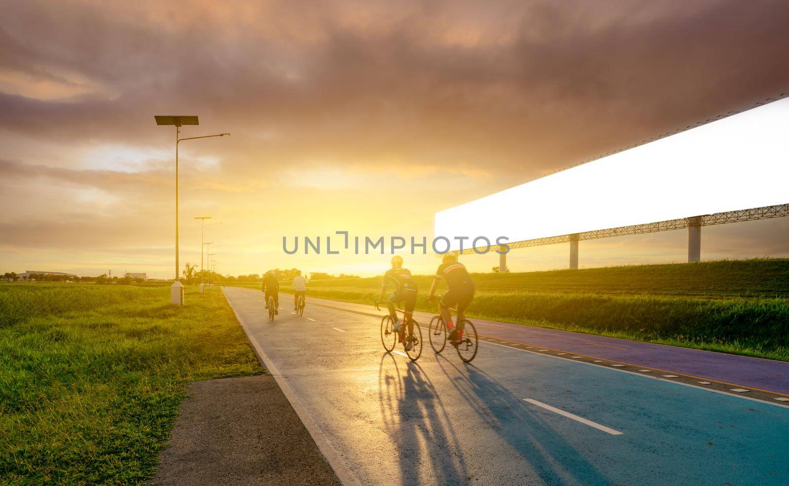 Sports man ride bicycles on the road in the evening near blank advertising billboard with sunset sky. Summer outdoor exercise for healthy and happy life. Cyclist riding mountain bike on bike lane.  by Fahroni
