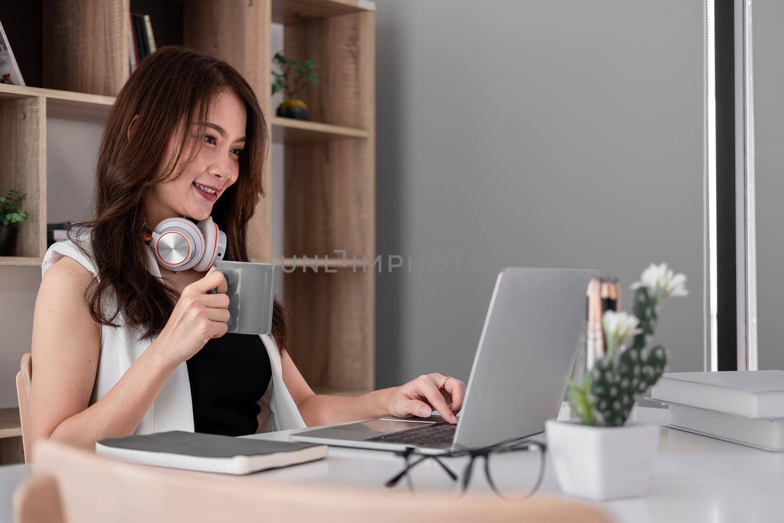 Asian woman with cup of coffee using laptop computer with headset at home, Online learning education concept by nateemee