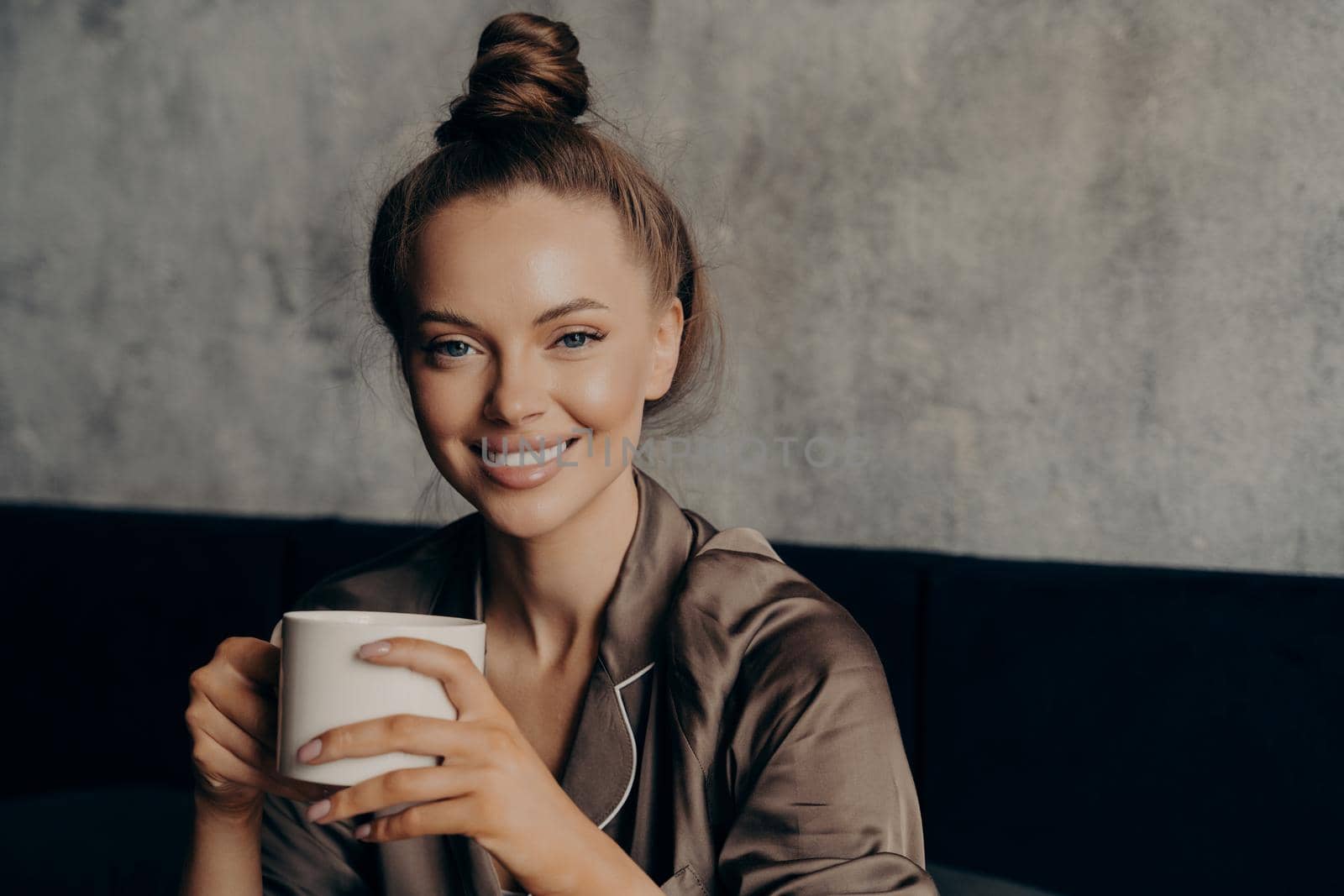 Young attractive cheerful woman with beautiful wide smile holding her morning cup of coffee and smiling at camera while sitting in bedroom in modern apartmnet after waking up, wears satin brown pajama