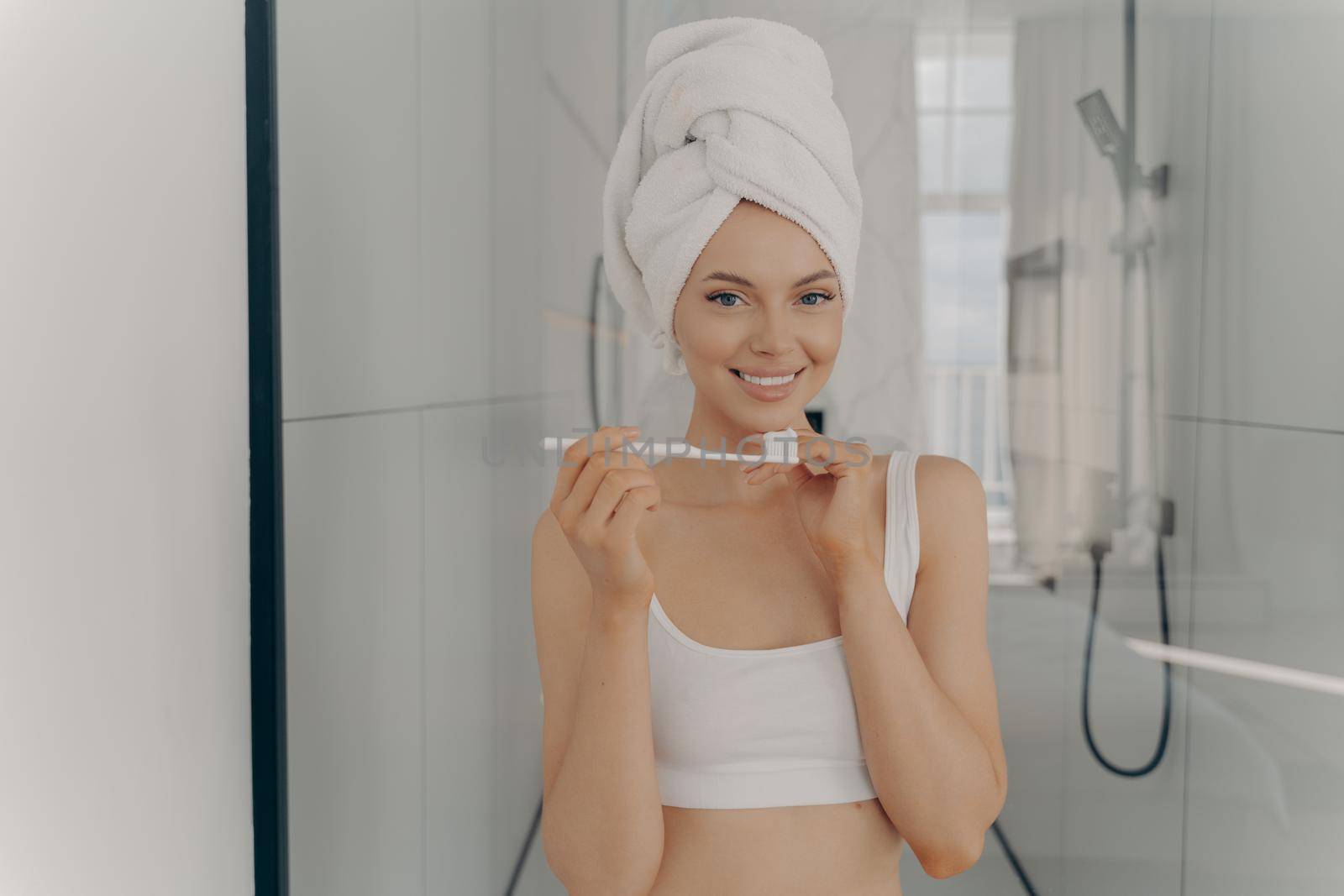 Happy beautiful lady with healthy perfect smile brushing teeth while standing in bathroom, wears towel wrapped on wet hair after morning shower. Personal body care and oral hygiene concept