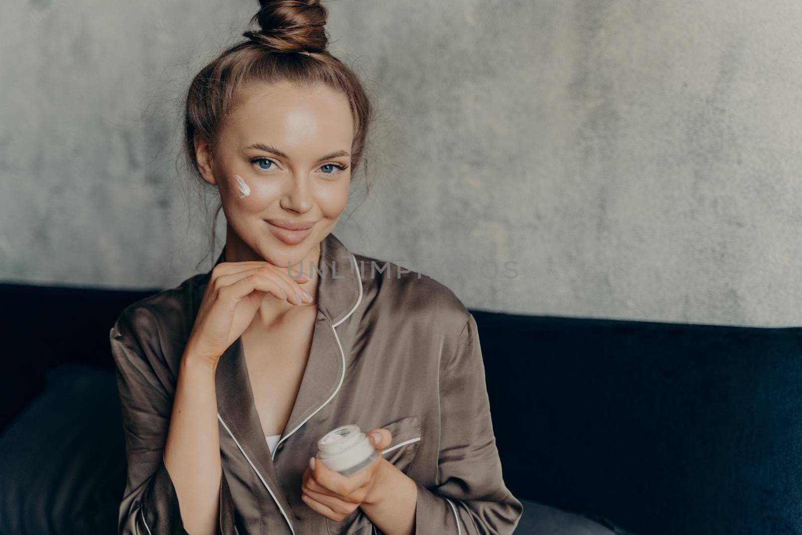 Close up shot of attractive young female model in brown silk pajama sitting on bed in morning after waking up with healthy fresh skin holding cream container in hand and applying cosmetic product