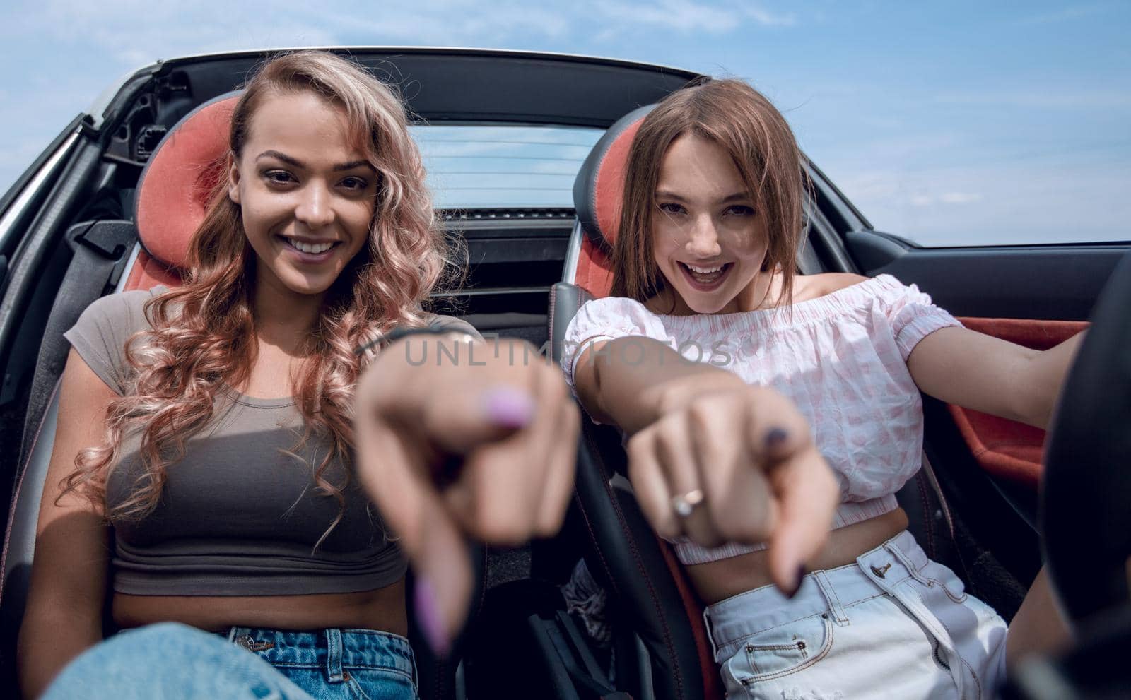 two happy young women sitting in a car and pointing at you. by asdf
