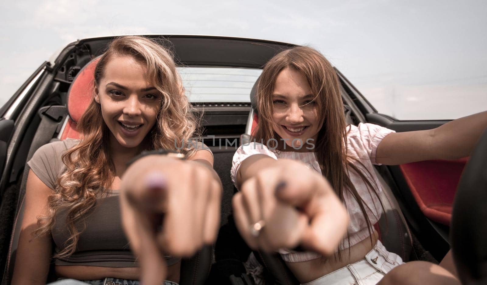 two happy girlfriends sitting in a car and pointing at you by asdf