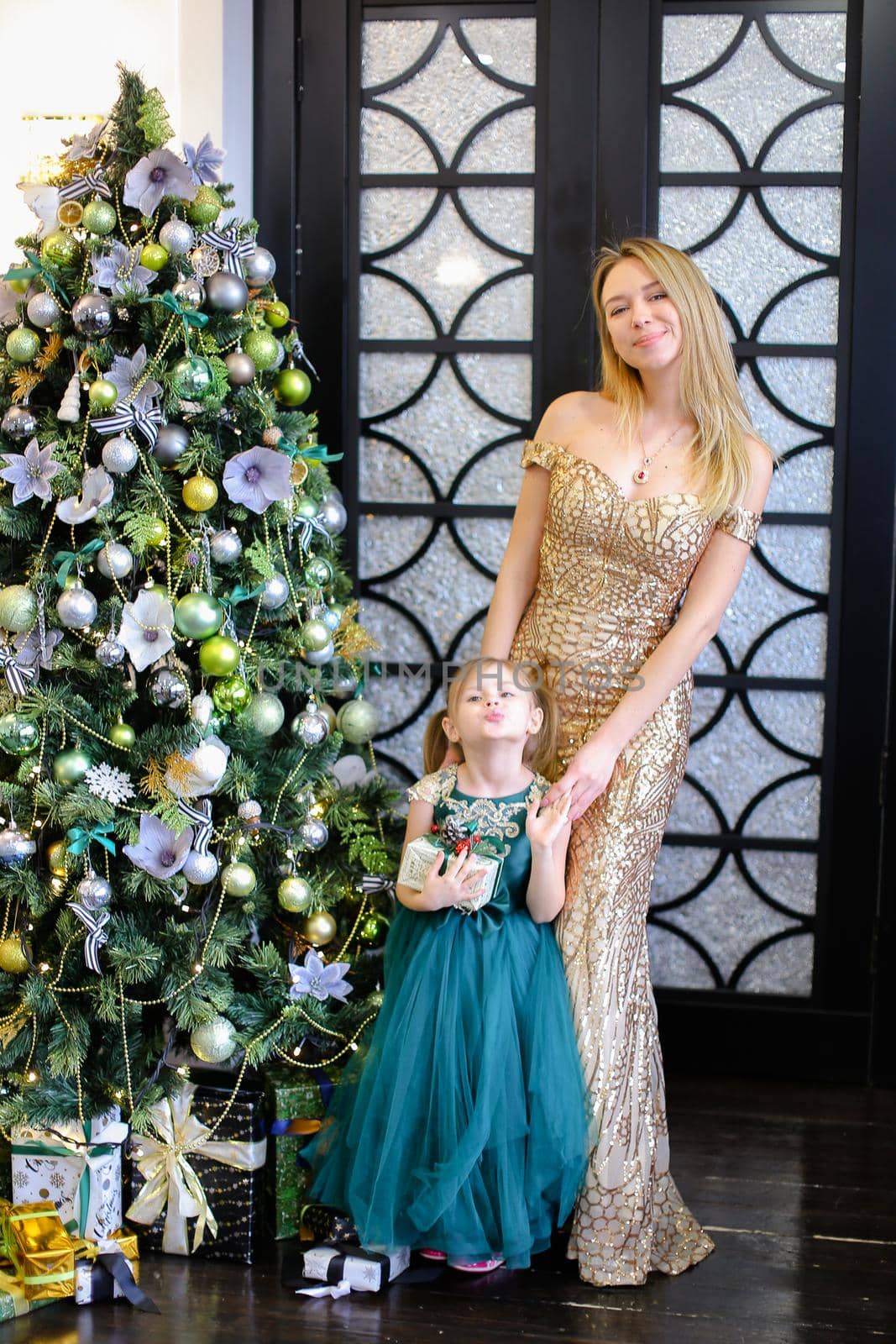 Young caucasian mother and little daughter wearing dresses standing near Christmas tree. by sisterspro