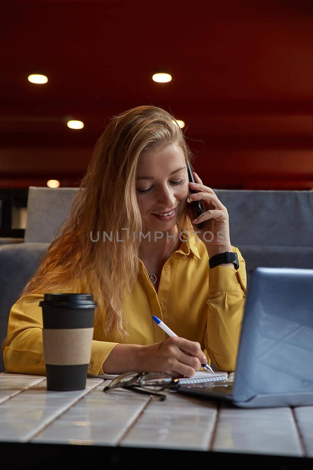 young well dressed cauasian woman sitting at table, calling by smartphone. beautiful lady talking by phone at cafe, making notes in notebook. modern communication technology, distant work, remote job
