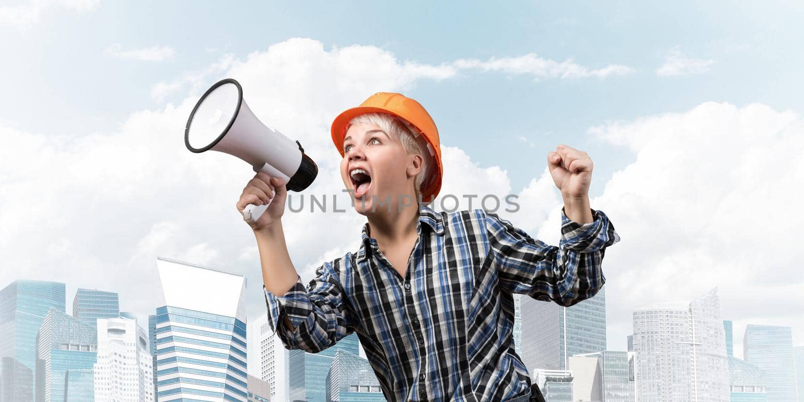 Expressive woman in safety helmet shouting into megaphone. Portrait of young emotional construction worker with loudspeaker on background of modern city. News announcement and advertisement.