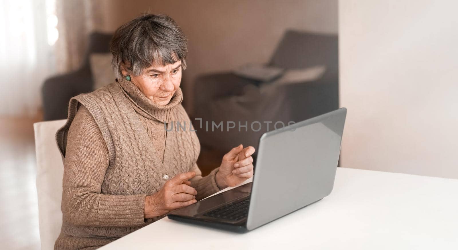 Old woman wearing casual clothes such as knitted sweater, spending free time at cozy home, sitting, resting using pc, typing message and searching, reading news online.