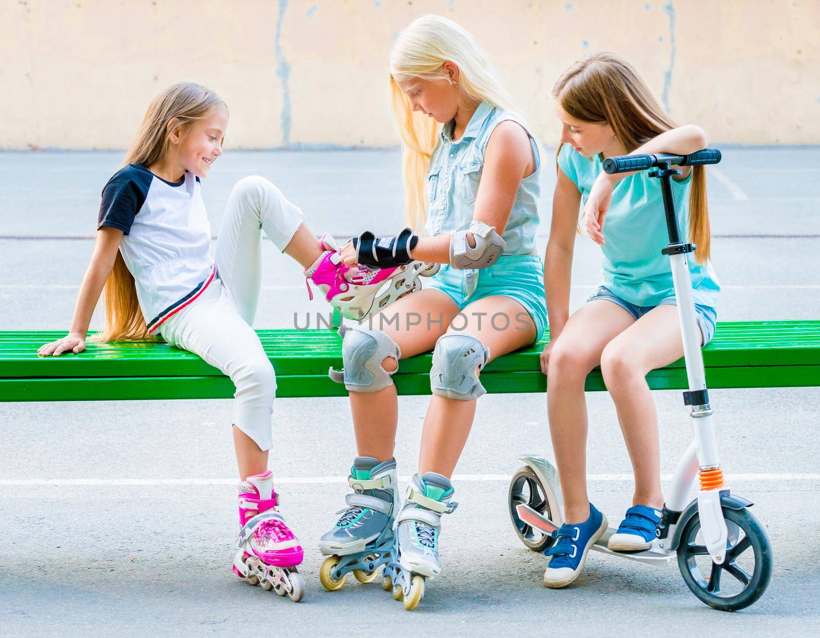 Little girls sitting on the green bench putting on rollerskates