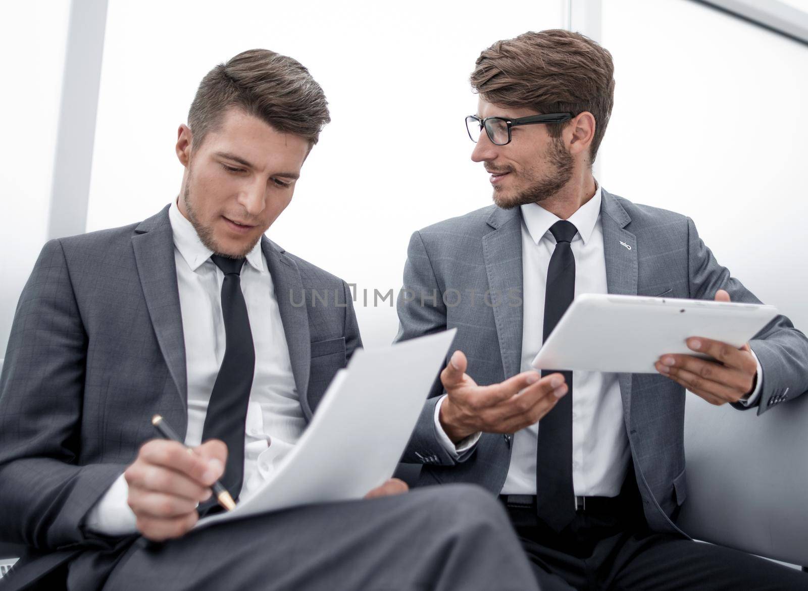 two young people are sitting in the office. One holds a tablet and gesticulates. The other holds a paper and a pen and listens.