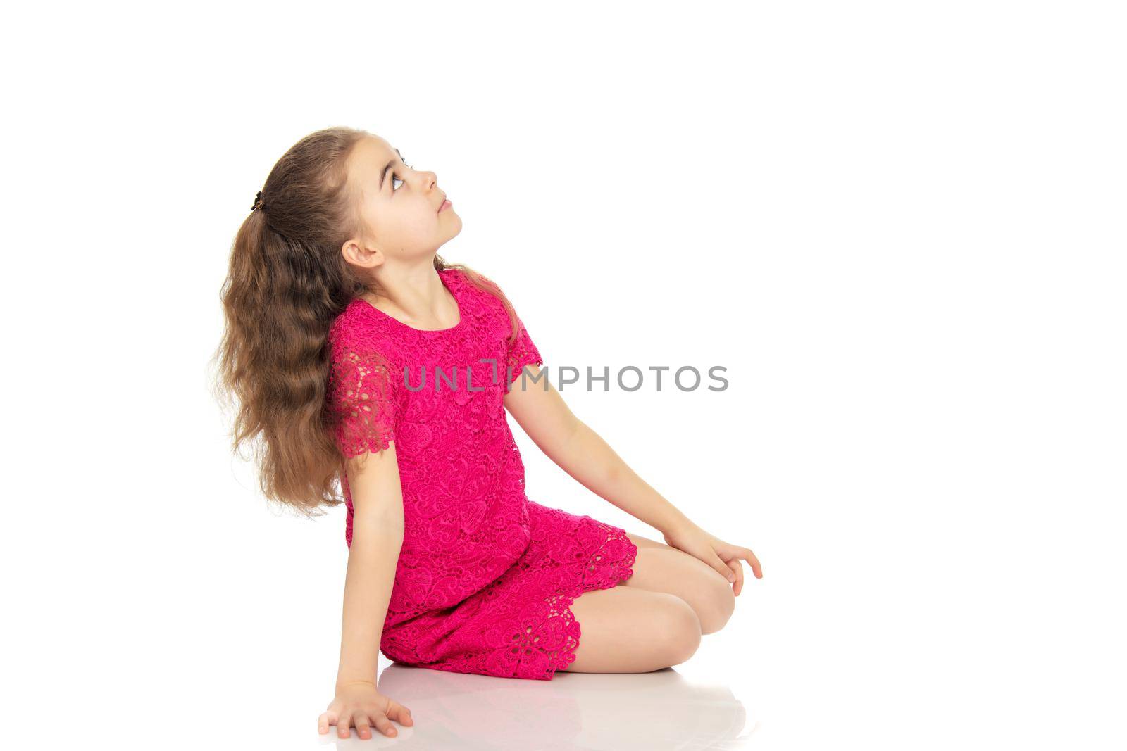 A beautiful little girl of primary school age, with long flowing hair, in a bright red dress.She sat down on her knees and looked up at the camera, turning sideways.Isolated on white background.