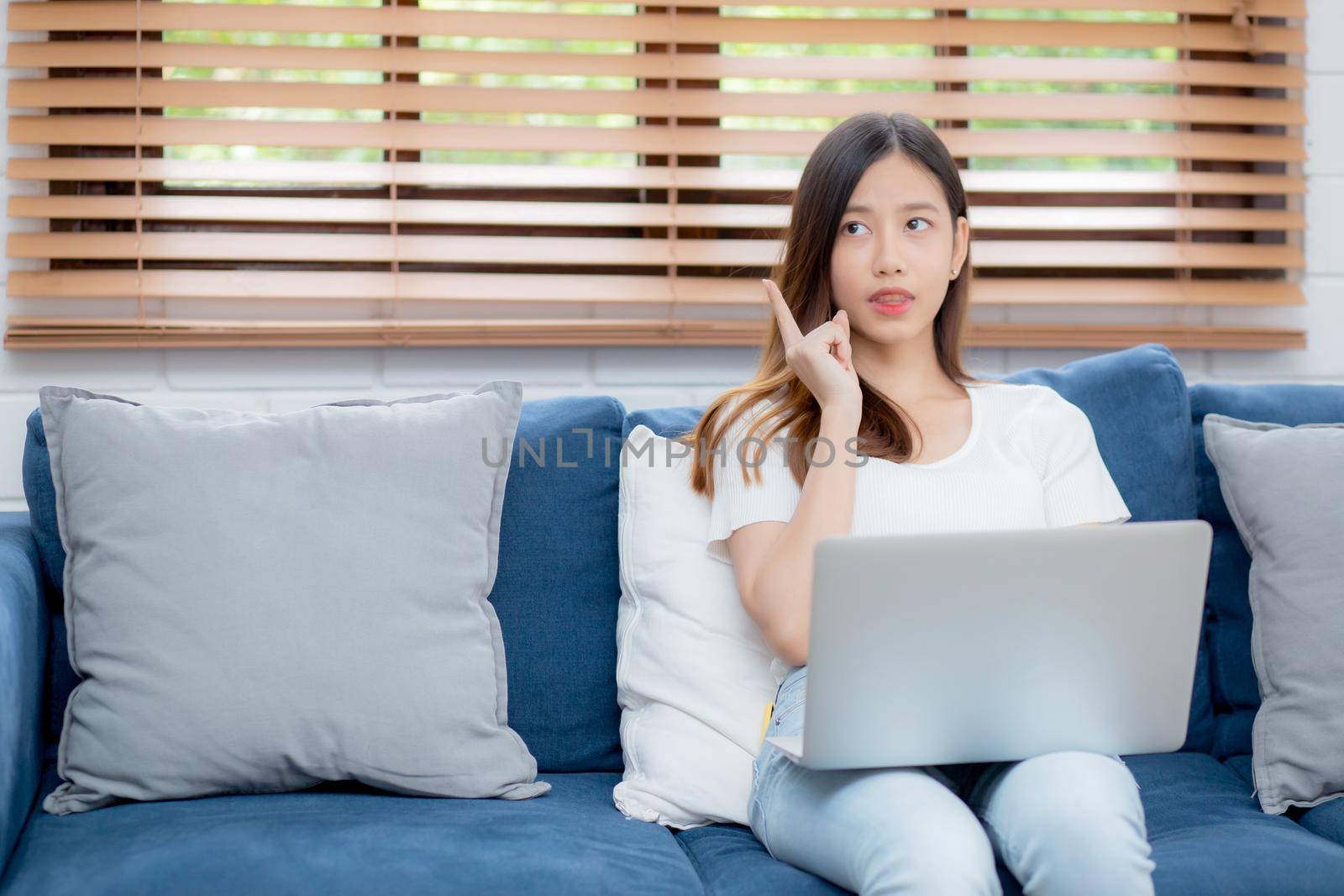 Beautiful young asian woman working on laptop computer and thinking idea on sofa at home, freelance girl sitting on couch using notebook to internet at living room, one person, lifestyle concept.