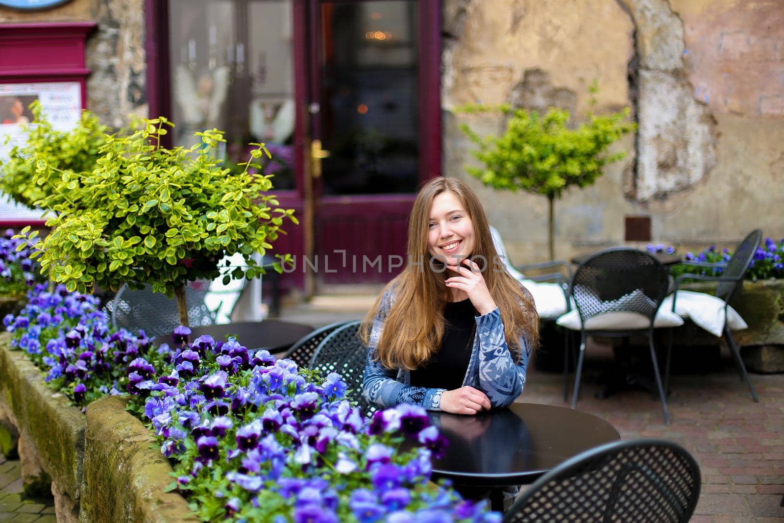 Young woman sitting at street cafe near blue flowers and green plants. Concept of cozy place in open air and resting.