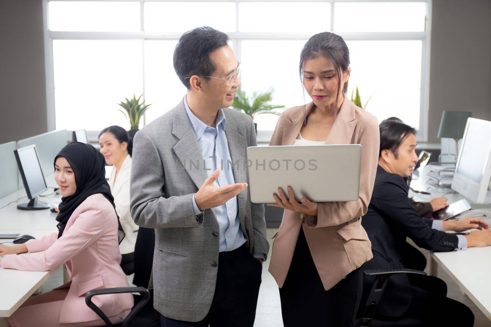 Asian businessman and businesswoman talking and looking laptop computer in the office, man standing explaining about financial with woman together, entrepreneur and employee planning about business. by nnudoo