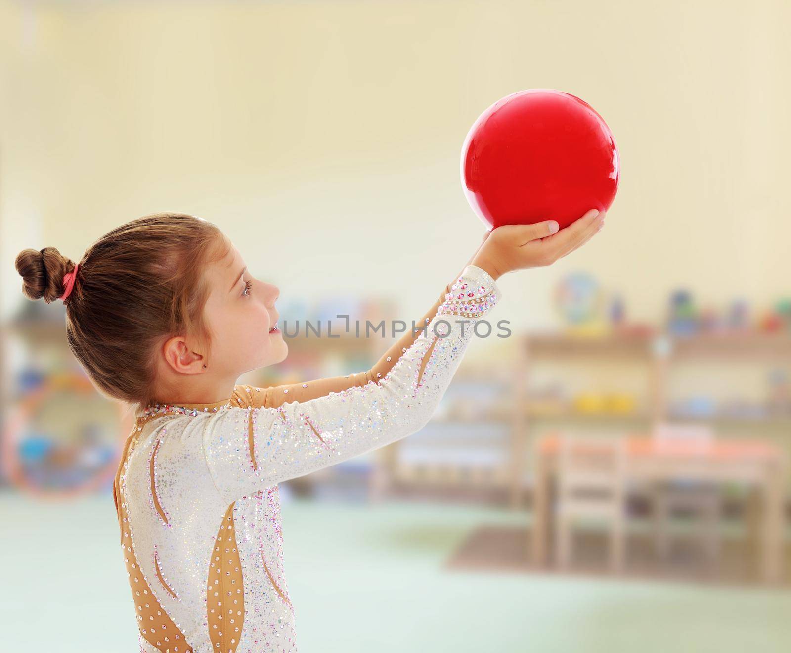 Cute little girl gymnast turned sideways to the camera , holding in his outstretched hands a red ball.On the background of the room where children live, and are on the shelves of toys.11