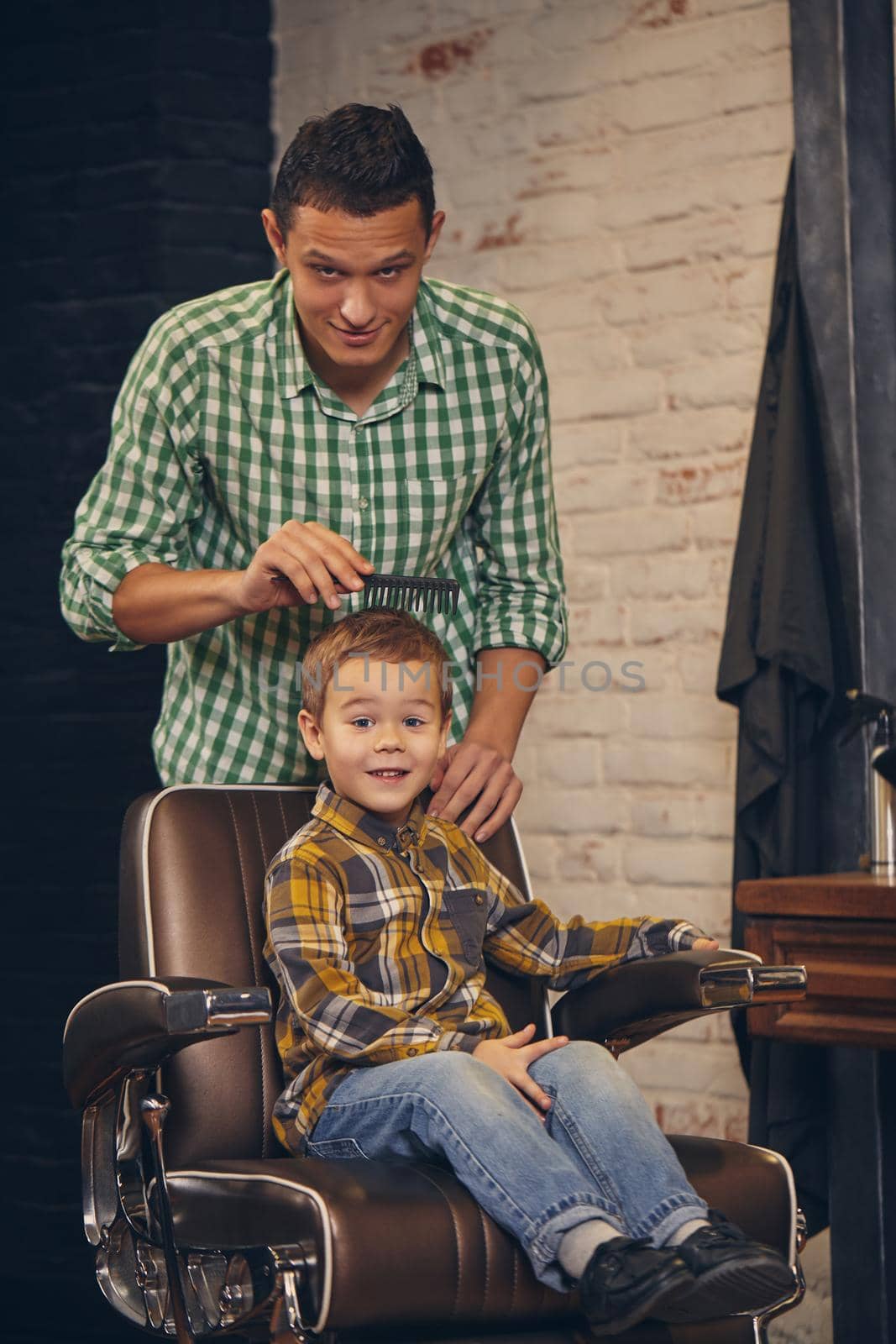 stylish little kid sitting on chair at barbershop with his young father on background, they are fooling around and having fun