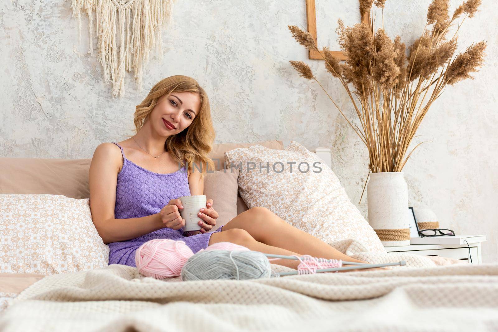 Young woman holding a mug with tea or coffee sitting on the bed in the bedroom. Near lie yarn and knitting needles. Horizontal photo. Freelance creative working and living concept.