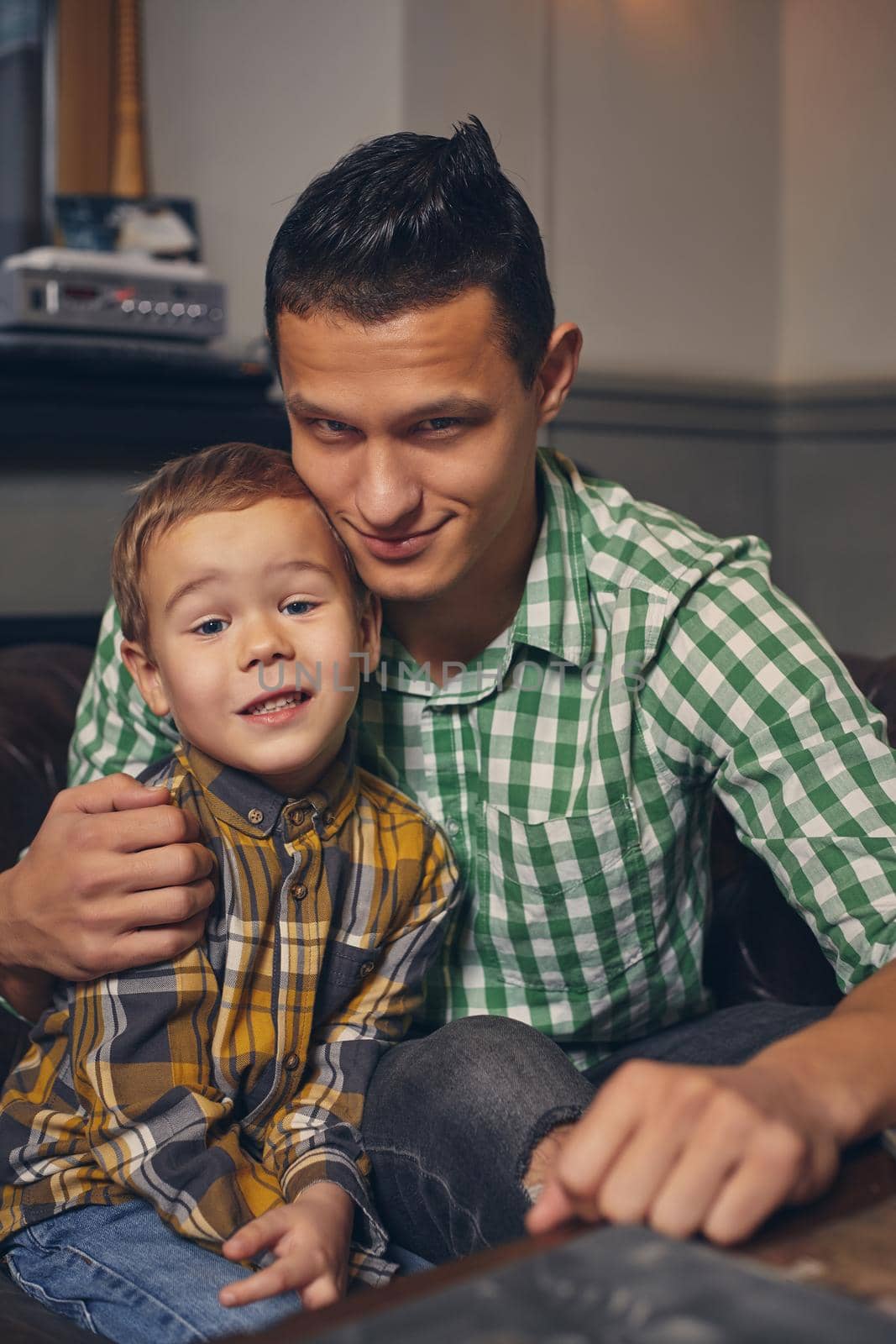 Young father and his stylish little son in the barbershop in the waiting room. They are waiting for the master, reading the fashion magazine and having a good time. Father hugs his son