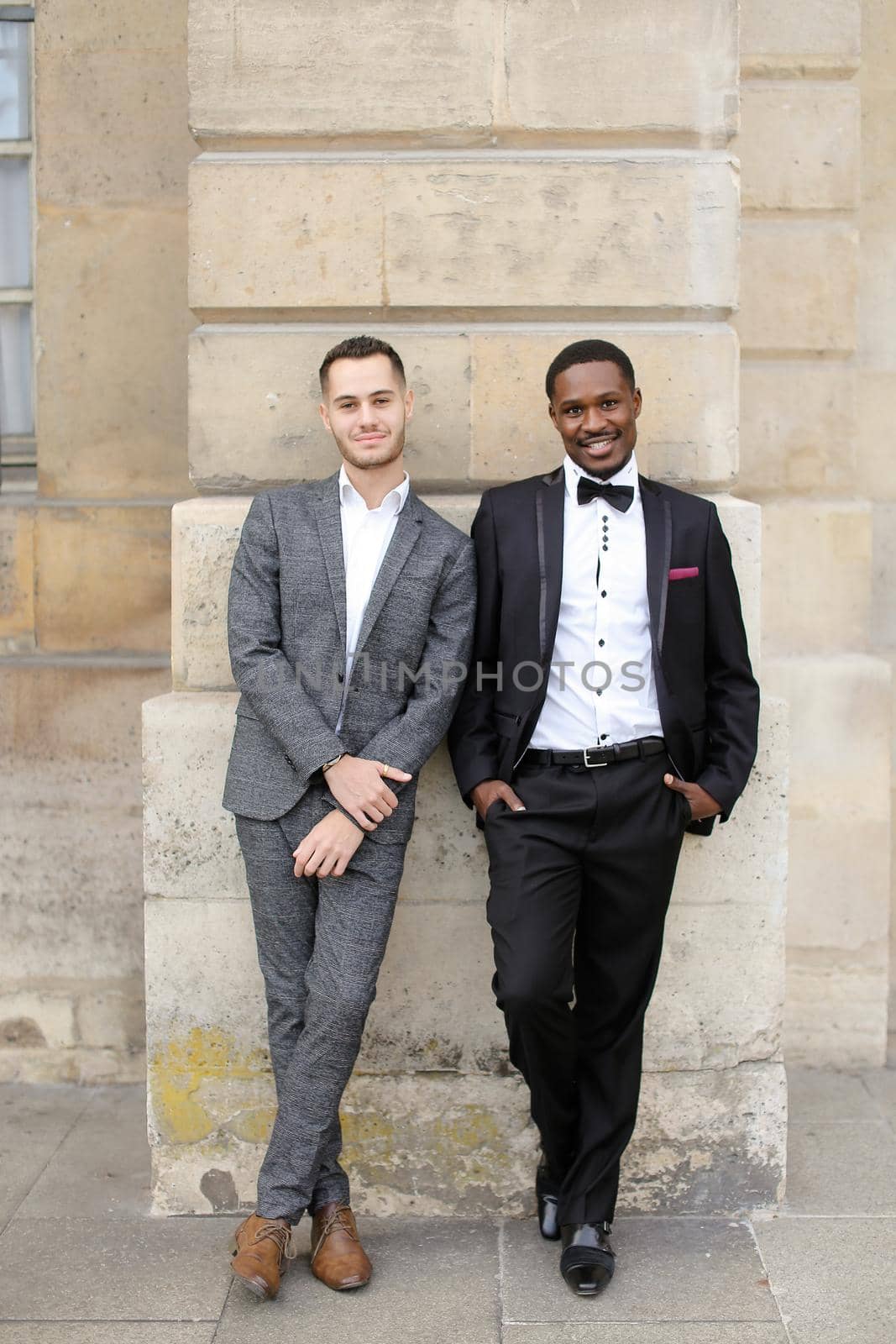 Afro american and caucasian happy gays standing near building and wearing suits. Concept of lgbt and walking in city.