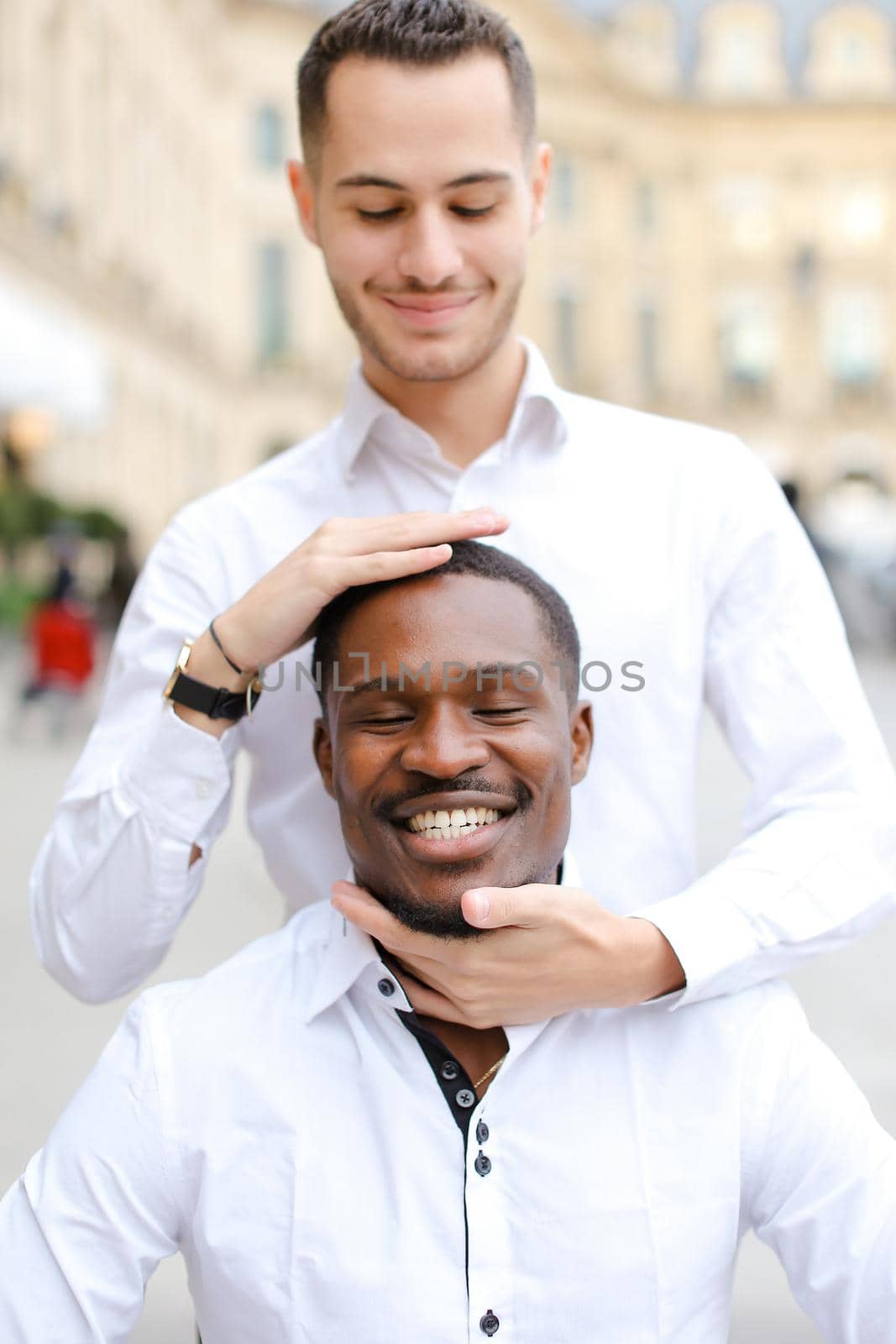 Caucasian boy holding afro american guy head by hands, wearing white shirt. Concept of stylish boy and haircut.