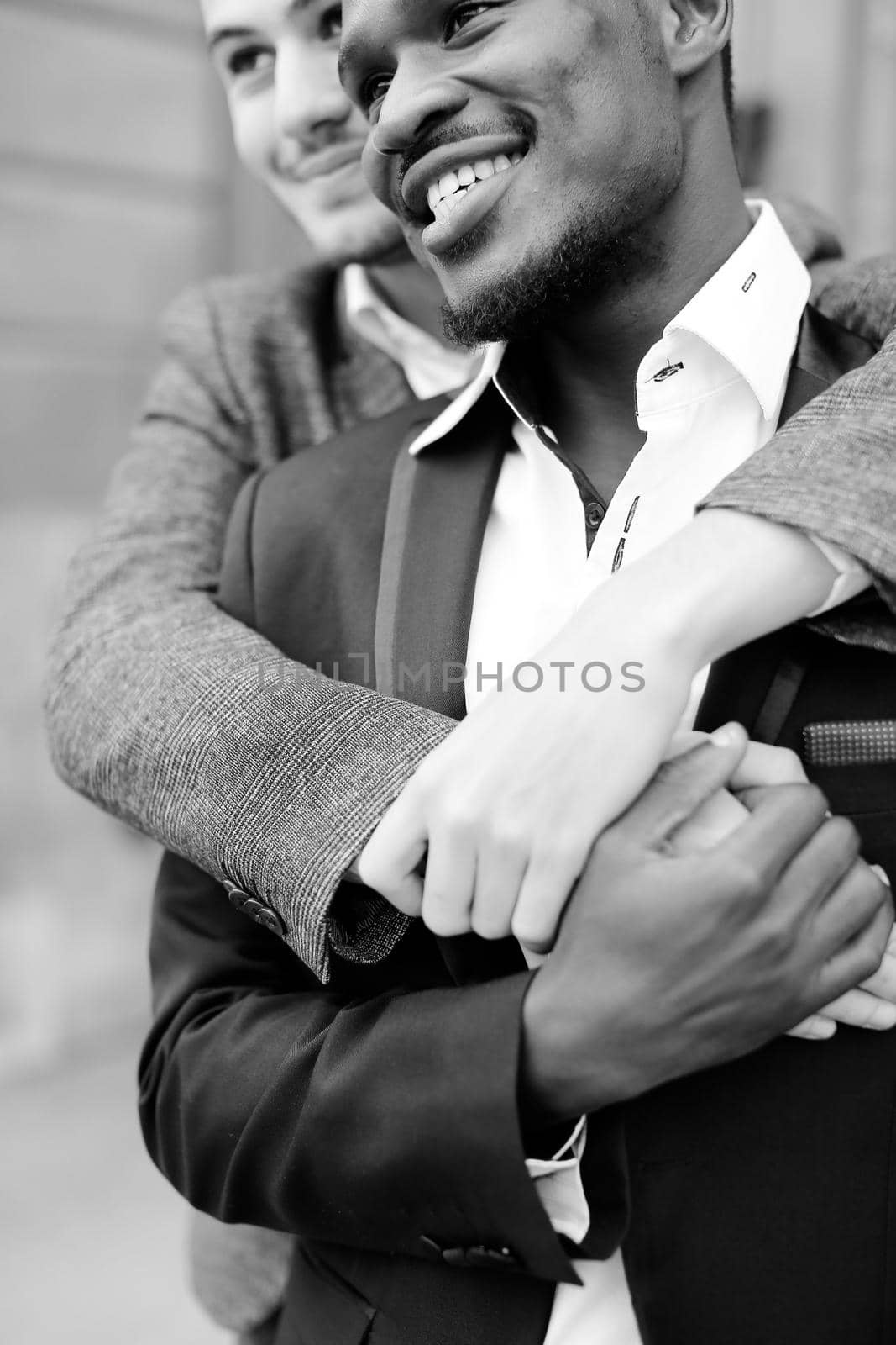 Black and white portrait of two hugging smiling boys, caucasian and afro american, wearing suits. Concept of gays and lgbt.