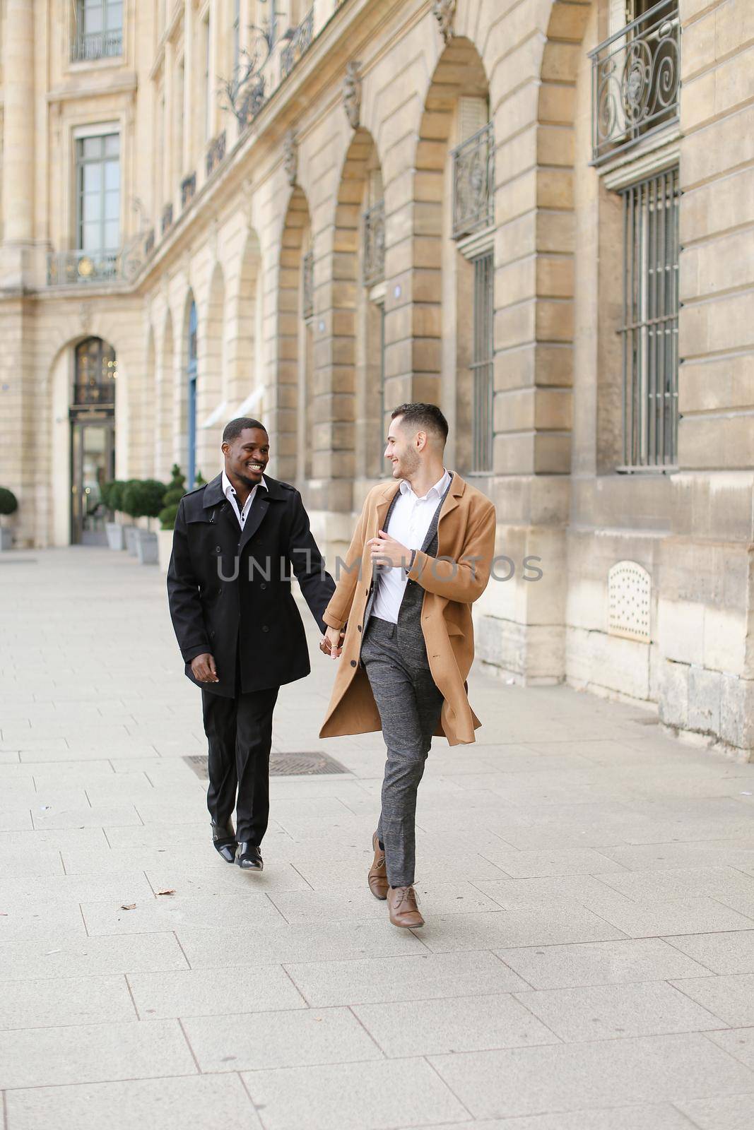 Caucasian man running with afroamerican male person and holding hands in city. Concept of happy gays and strolling.