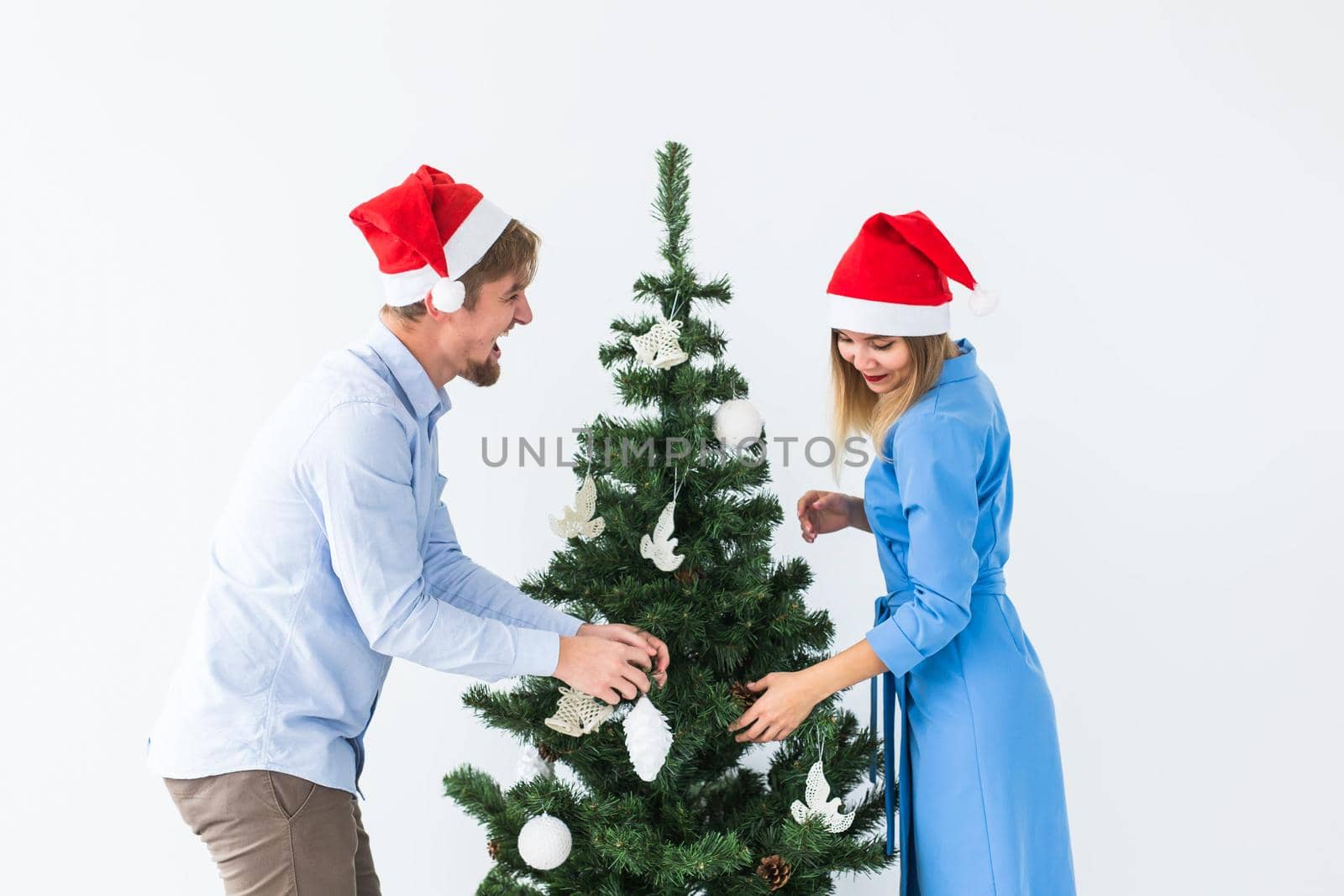 Holidays and festive concept - Young family couple in santa hats decorating the christmas tree by Satura86