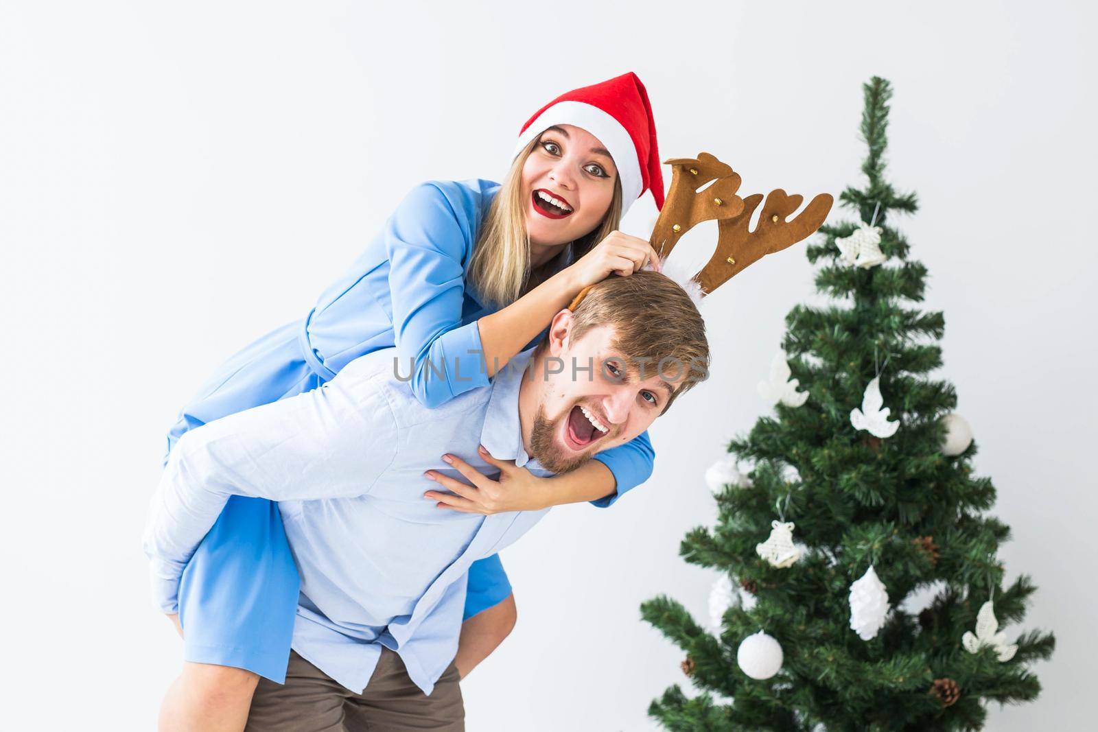Funny man giving piggyback to his wife while they wearing Santa hats for Christmas holidays at home. by Satura86