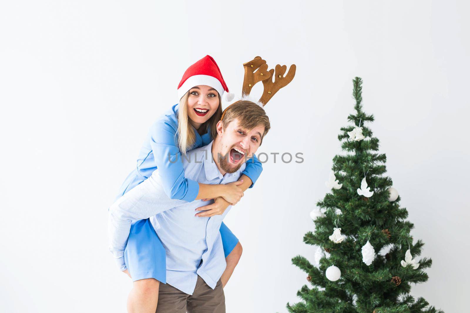 Funny man giving piggyback to his wife while they wearing Santa hats for Christmas holidays at home. by Satura86