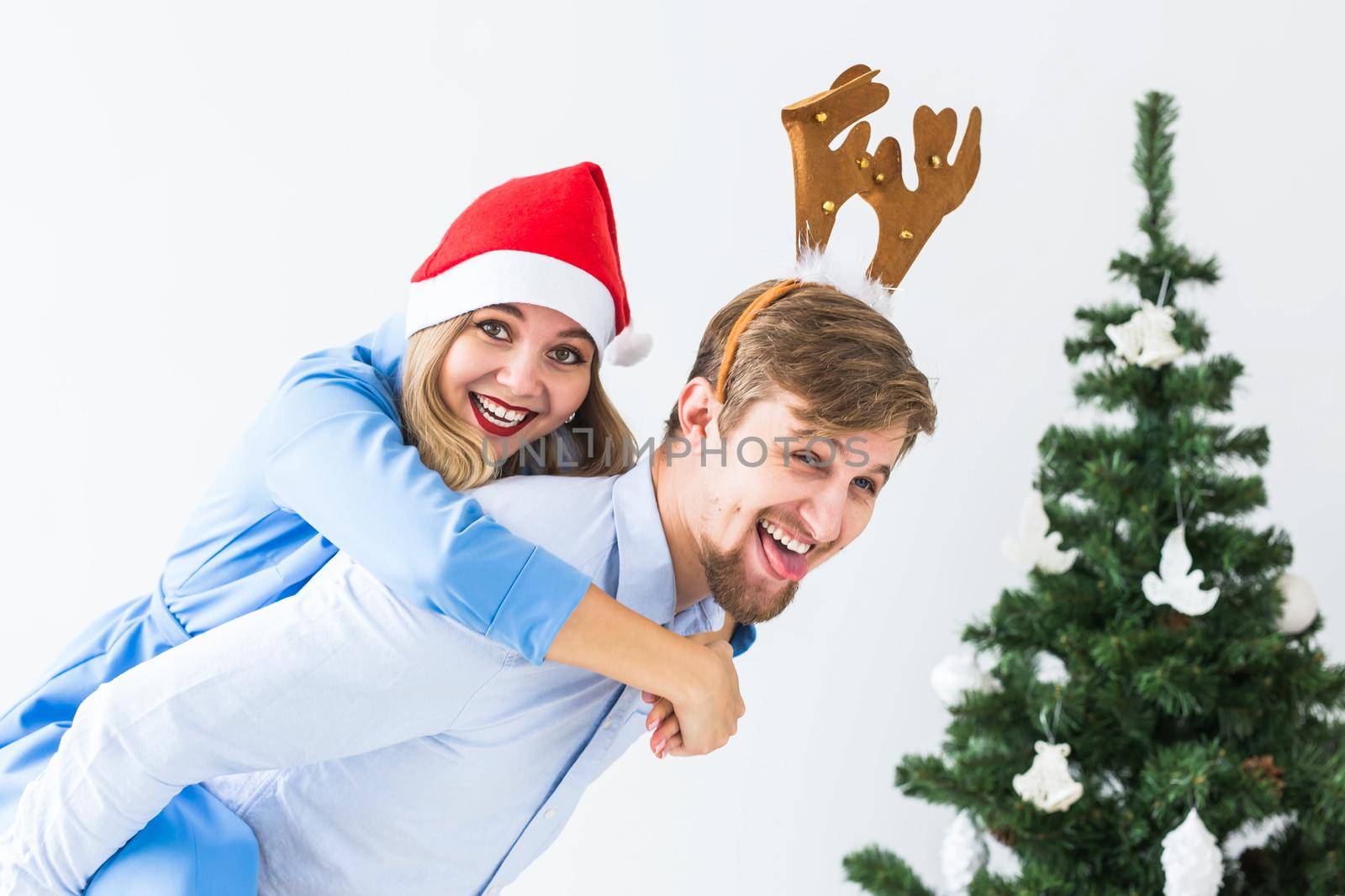 Funny man giving piggyback to his wife while they wearing Santa hats for Christmas holidays at home. by Satura86