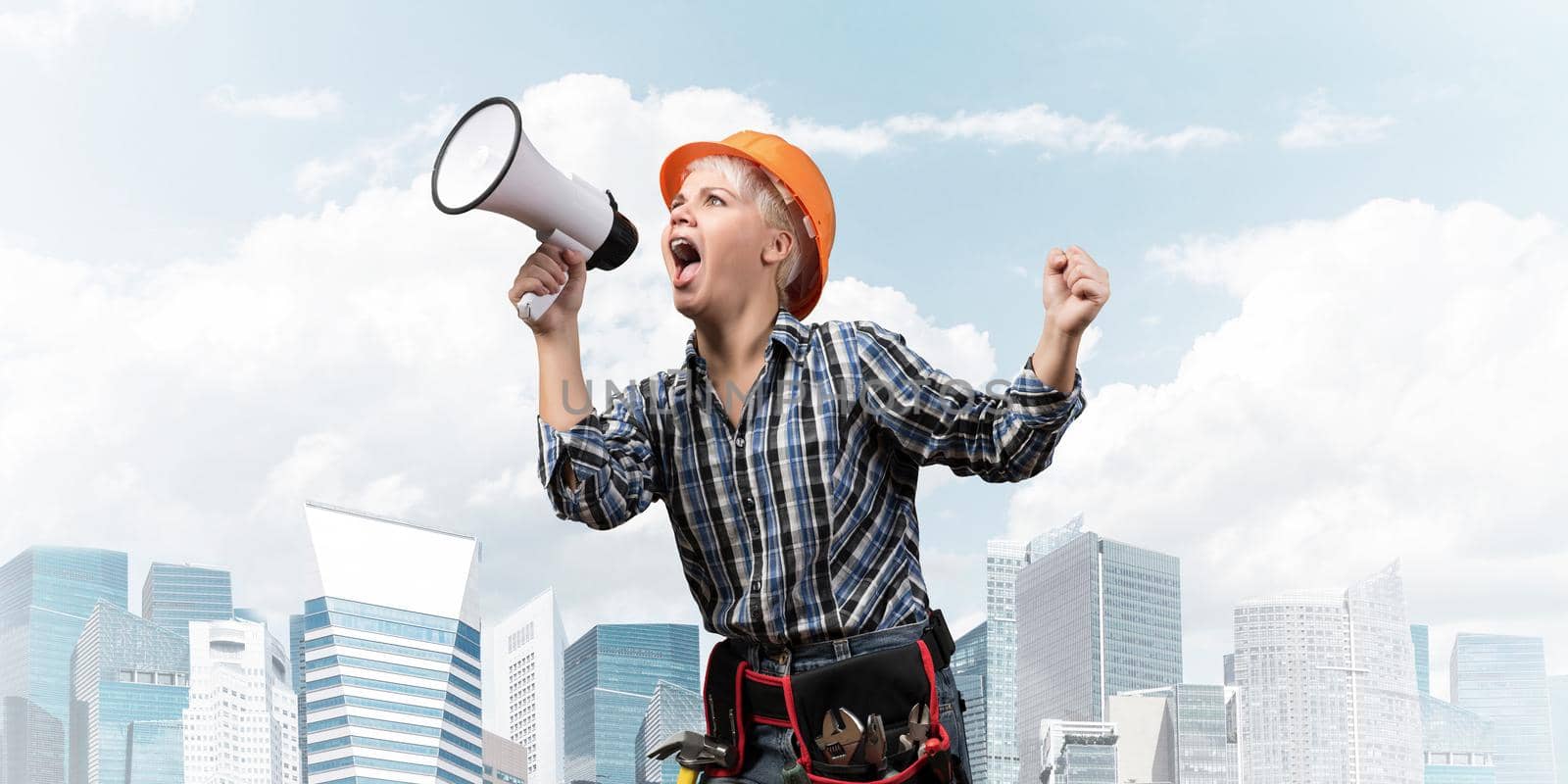Expressive woman in safety helmet shouting into megaphone. Portrait of young emotional construction worker with loudspeaker on background of modern city. News announcement and advertisement.