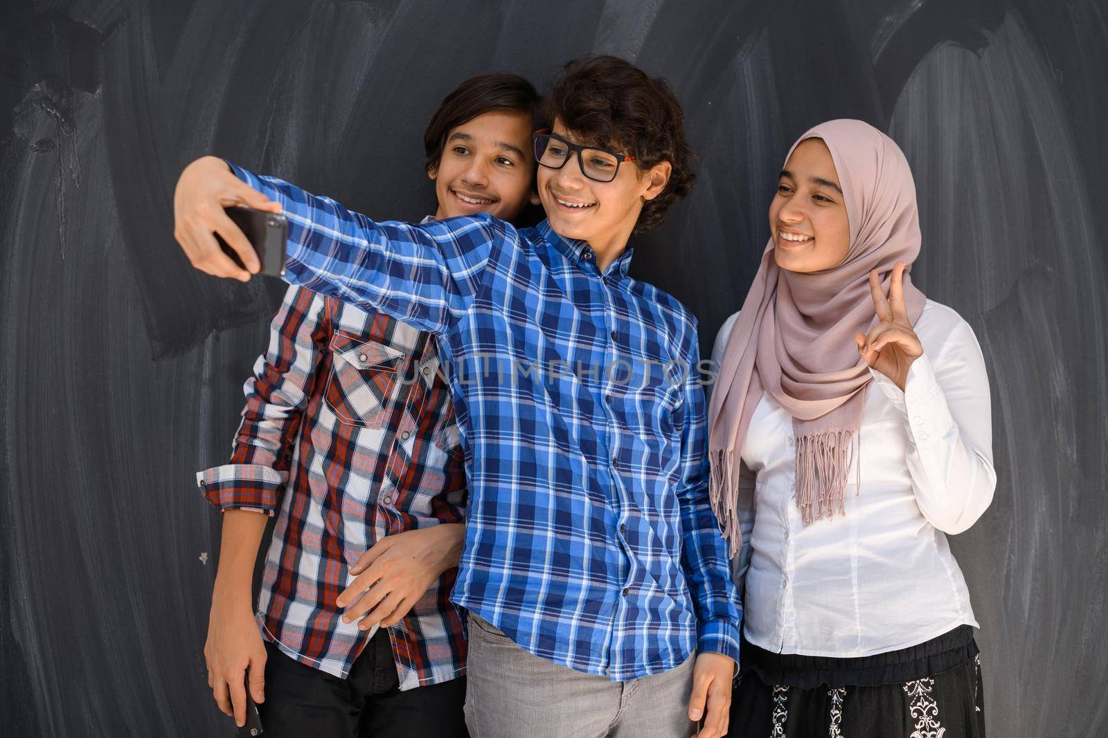 Group of Arab teens taking selfie photos on a smartphone with black chalkboard in the background. Selective focus. High quality photo